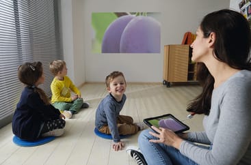 Eine Frau hält ein Tablet in der Hand und sitzt mit drei kleinen Kindern auf dem Boden. Sie sehen ein Bild von zwei Pflaumen, das auf eine weiße Wand in einem hellen Klassenzimmer projiziert wird.