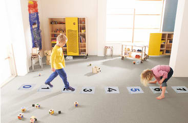 Zwei Kinder spielen drinnen auf nummerierten Matten Himmel und Hölle. Der Junge in Gelb tritt auf ein Quadrat, während das Mädchen in Rosa sich in der Nähe der Zahlen bückt. In dem hell erleuchteten Raum sind Spielzeuge und Regale zu sehen.