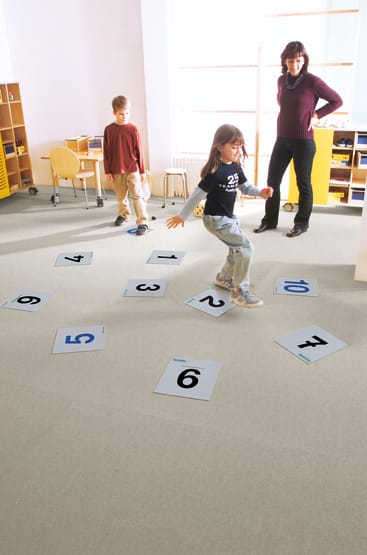 Zwei Kinder und ein Erwachsener befinden sich in einem Klassenzimmer. Ein Kind schreitet zwischen nummerierten Matten auf dem Boden, während das andere Kind und der Erwachsene zusehen. Im Hintergrund sind Regale und Stühle zu sehen.