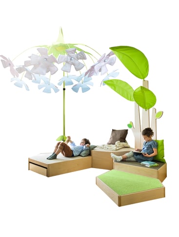 Zwei kleine Kinder sitzen auf modularen Holzmöbeln mit grünen Kissen, neben übergroßen dekorativen Blättern und einer großen schirmähnlichen Struktur mit weißen und grünen Blütenblättern, vor einem weißen Hintergrund.