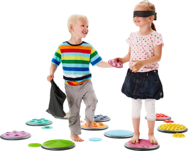 Ein Junge und ein Mädchen mit verbundenen Augen stehen auf strukturierten Trittsteinen. Der Junge hält einen Stoffbeutel und führt das Mädchen an der Hand. Beide Kinder lächeln und sind barfuß. Der Boden ist mit verschiedenen bunten Scheiben übersät.