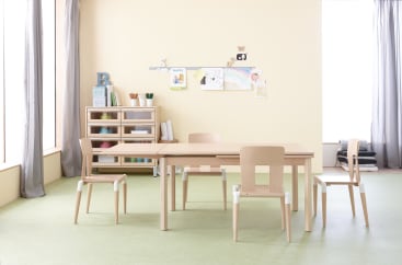 Ein Klassenzimmer mit vier Holzstühlen und einem rechteckigen Tisch, Regalen mit Büchern und Material und bunten Kinderzeichnungen an einer hellgelben Wand. Große Fenster lassen natürliches Licht herein.