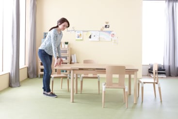 Eine Frau in Freizeitkleidung stellt leere Holzstühle um einen hellen Holztisch in einem hellen Klassenzimmer mit Zeichnungen an der Wand und Regalen im Hintergrund auf.