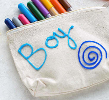 Ein beiges Stoffmäppchen mit dem Wort "Boy" und einer mit blauer Puff-Farbe gezeichneten Spirale, das mehrere bunte Marker enthält, die oben teilweise sichtbar sind.