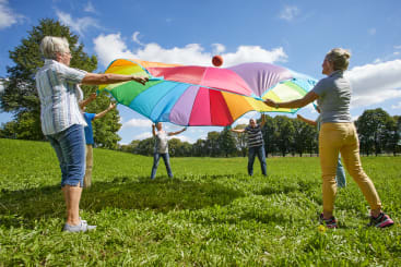 Eine Gruppe Erwachsener steht auf einer Wiese und hält die Ränder eines großen, bunten Fallschirms fest. Sie heben den Fallschirm an und schicken einen roten Ball in die Luft. Im Hintergrund sind Bäume und ein blauer Himmel mit Wolken zu sehen.