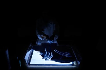 Eine Person in einem Laborkittel steht in einem dunklen Raum und zeichnet mit einem dünnen Werkzeug Muster in eine mit dunklem Granulat gefüllte Schale, die von unten durch eine helle rechteckige Lichtquelle beleuchtet wird.