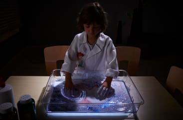 Ein Kind, das einen weißen Laborkittel trägt, zeichnet mit beiden Händen Muster in ein Tablett, das mit Sand und Glitter gefüllt ist und auf einem Tisch steht. Der Raum ist schwach beleuchtet und das Tablett wird von unten angestrahlt.