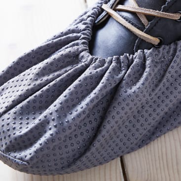Nahaufnahme eines Fußes, der einen dunklen Schuh trägt, der von einer grauen, strukturierten und gerafften Schutzhülle aus Stoff mit kleinen Punktmustern bedeckt ist, die auf einer hellen Holzfläche ruht.