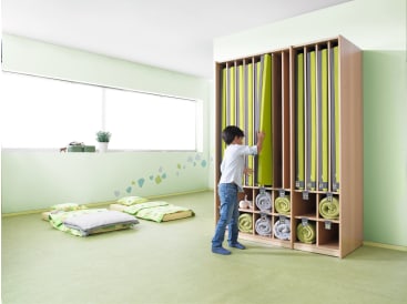 In einem hellen, minimalistischen Raum mit grünen Wänden und verstreuten Kissen auf dem Boden zieht ein kleines Kind eine grüne Matte aus einem hohen Holzregal mit aufgerollter Bettwäsche und Decken heraus.