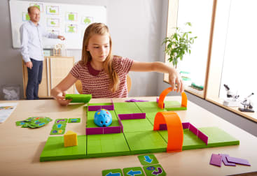 Ein Mädchen bewegt Figuren auf einem grünen und lilafarbenen Labyrinthspiel mit einem blauen Ball auf einem Tisch, während ein Erwachsener in einem Klassenzimmer mit großen Fenstern und sichtbaren Unterrichtsmaterialien neben einer Tafel steht.