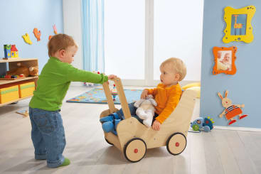 Zwei Kleinkinder befinden sich in einem hell erleuchteten Spielzimmer. Ein Kind steht und schiebt einen hölzernen Spielzeugwagen, während ein anderes Kind darin sitzt und Stofftiere hält. Das Zimmer ist mit Spielzeug, bunten Dekorationen und Fotos an der Wand ausgestattet.