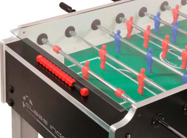 Nahaufnahme eines Kickertisches mit roten und blauen Spielerfiguren, Metallstangen mit roten Griffen, einem grünen Spielfeld und einer Glasabdeckung. Ein Teil des schwarzen Rahmens und des Logos des Tisches sind sichtbar.