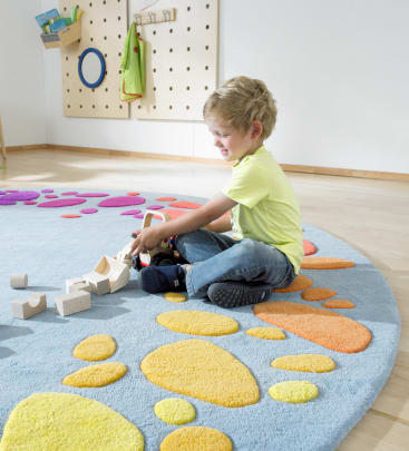 Ein kleines Kind sitzt auf einem blauen Teppich mit orangefarbenen und gelben Formen und spielt mit Holzklötzen. Der Raum hat einen hellen Holzboden und eine Wand mit Haken, an denen kleine Gegenstände hängen.