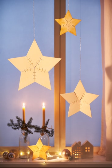 Vor einem Fenster hängen drei hölzerne Stern-Ornamente. Darunter befinden sich zwei brennende Kerzen in Glasflaschen, ein kleiner Fichtenzweig, dekorative Lichter, Weihnachtsschmuck und weiße, hausförmige Schachteln.