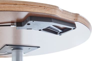Nahaufnahme der Unterseite einer runden Holztischplatte, die einen schwarzen Mechanismus, der an der Oberfläche befestigt ist, und eine Stützstange aus Metall zeigt. Die geschichtete Kante des Holzes ist sichtbar.