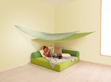 Ein Kind liegt auf einem grünen Bettsofa mit mehreren Kissen und einer grauen Decke und hält ein Stofftier. Über dem Sofa ist ein großer grüner Stoffhimmel angebracht, der einen zeltartigen Schutz in einem minimalistischen, hellen Raum schafft.