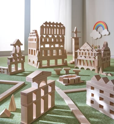 Holzbauklötze sind auf einer grünen Fläche zu verschiedenen Strukturen angeordnet. Im Hintergrund befinden sich ein weißer Vorhang und eine Wanddekoration in Form eines Regenbogens mit Wolken und Regentropfen.