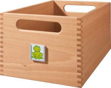 Eine Aufbewahrungsbox aus hellem Holz mit rechteckig ausgeschnittenen Griffen und einem kleinen quadratischen Etikett mit einem grünen Cartoon-Frosch auf der Vorderseite.