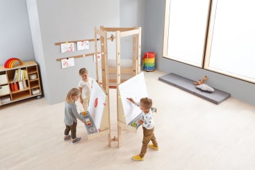 In einem hellen Klassenzimmer mit hellem Holzfußboden und Regalen mit Malutensilien malen drei kleine Kinder auf großen Papierbögen, die an einer Holzstaffelei befestigt sind. An der Wand hinter ihnen sind Zeichnungen ausgestellt.