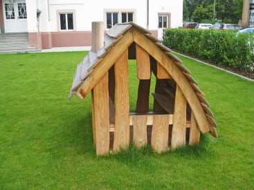 Ein kleines, rustikales Holzspielhaus mit einem schrägen Dach steht auf einer grünen Wiese in der Nähe eines Gebäudes. Die Struktur besteht aus unebenen Holzbrettern und hat offene Seiten und eine Bank im Inneren.