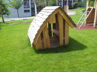 Ein kleines hölzernes Spielhaus mit einem schrägen Dach steht auf einer Rasenfläche auf einem Spielplatz. Im Hintergrund sind Bäume, Spielgeräte und ein weißes Gebäude zu sehen.