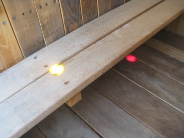 Eine Holzbank mit einem gelben Lichtpunkt auf der Sitzfläche und einem roten Lichtpunkt auf dem Boden darunter, wobei beide Lichter durch schmale Spalten zwischen den Holzbrettern scheinen.