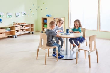 Drei kleine Kinder sitzen in einem hellen, aufgeräumten Klassenzimmer um einen runden Tisch herum und zeichnen und malen. Im Hintergrund sind Holzstühle und Regale mit Unterrichtsmaterialien und Spielzeug zu sehen.