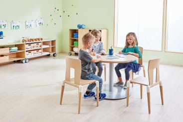 Drei kleine Kinder sitzen um einen runden Tisch in einem hellen Klassenzimmer und zeichnen oder schreiben auf Papier. Im Hintergrund sind Regale mit Material und Dekoration zu sehen. Der Raum hat helle Holzböden und große Fenster.