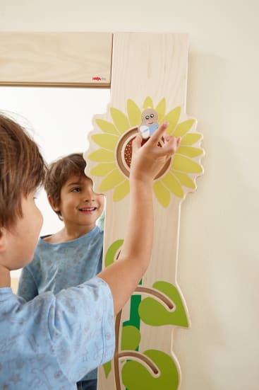 Ein Kind bewegt eine kleine Holzbiene entlang einer Sonnenblumenbahn, die an der Seite eines Spiegels befestigt ist. Das Kind lächelt und betrachtet sein Spiegelbild.