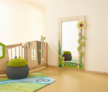 Eine Spielzimmerecke mit einem grün-blauen Teppich, einem runden grau-grünen Hocker, einem Ganzkörperspiegel mit Sonnenblumenrahmen und einem hölzernen Spielzaun mit Dekorationen zum Thema Natur.