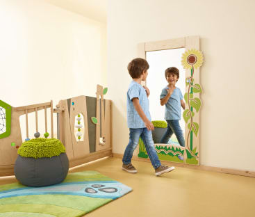 Ein kleines Kind steht vor einem an der Wand befestigten Spiegel, der mit einer Sonnenblume verziert ist, und betrachtet sein Spiegelbild. Das Zimmer hat einen hellen Fußboden, einen grün-blauen Teppich, einen weichen Sitzsack und Spielplatten aus Holz.
