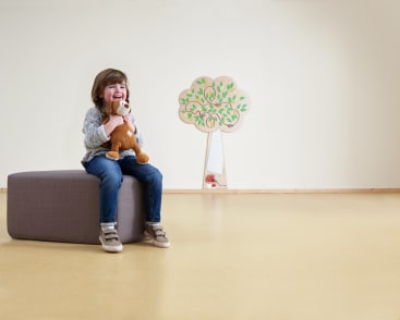 Ein kleines Kind sitzt auf einer grauen rechteckigen Bank, lächelt und umarmt ein Stofftier in einem geräumigen Zimmer mit einem hellgelben Boden und einem dekorativen Baum an der Wand im Hintergrund.