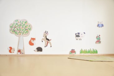 Ein minimalistisches Zimmer mit Wanddekorationen zum Thema Tiere und Natur, darunter ein Baum, ein Fuchs, ein Igel, ein Reh, eine Kuh, eine Eule und ein Marienkäfer. Der Boden ist beige mit einem grünen Teppich in der unteren rechten Ecke.