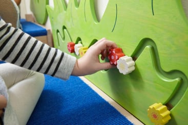 Ein Kind in einem gestreiften langärmeligen Hemd bewegt einen roten, blumenförmigen Block entlang einer grünen Wandtafel mit gewellten Spuren auf einem blauen Teppich.