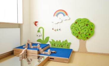 Ein Spielbereich für Kinder mit blauen Plattformen, auf denen Holzbausteine stehen, einem großen Spiegel und Wanddekorationen wie einem grünen Baum, einem Regenbogen mit Wolken, Gras, einer Blume und einem Marienkäfer.