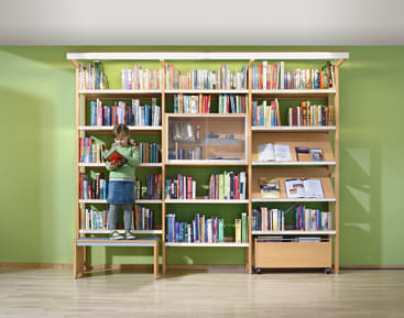 Ein kleines Kind steht auf einer Bank vor einem großen, mit bunten Büchern gefüllten Bücherregal und liest in einem roten Buch. Die Wand hinter dem Bücherregal ist hellgrün gestrichen. Der Boden ist aus hellem Holz.