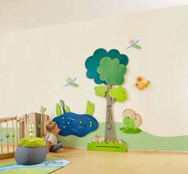 Ein kleines Kind sitzt auf dem Boden neben einer Wand, die mit einem Baum, einem Teich, einem Frosch, Libellen und Gras dekoriert ist, alles aus bunten Holzteilen in einer spielerischen, naturnahen Umgebung.