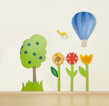 Eine mit bunten Holzschnitzereien verzierte Wand mit einem Baum, drei Blumen, einem Vogel und einem blauen Heißluftballon über einem beigen Boden.