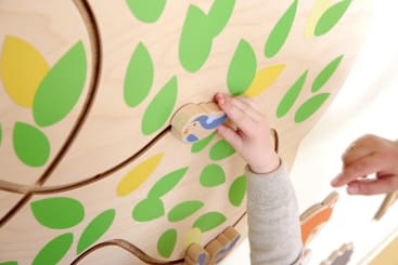 Die Hand eines Kindes bewegt eine kleine Holzfigur mit einer Vogelillustration entlang einer Bahn auf einem mit grünen und gelben Blättern verzierten Holzbrett. In der Nähe ist die Hand eines Erwachsenen zu sehen.