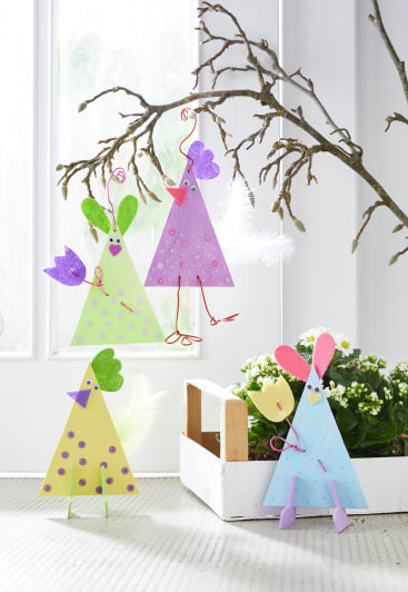Osterhühner und -vögel aus Papier in Pastellfarben hängen an einem kahlen Baumzweig und sitzen dort. Im Hintergrund stehen ein weißer Holzkasten mit Topfpflanzen und eine Glastür.