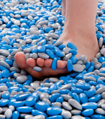 Nahaufnahme eines nackten Fußes, der teilweise von einer Mischung aus blauen und grauen ovalen Pralinen oder Bonbons bedeckt ist, wobei weitere Bonbons unter und um den Fuß verstreut sind.