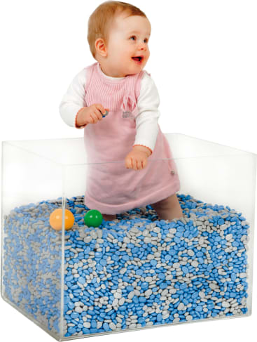 Ein Baby in einem rosa Kleid steht in einer durchsichtigen Schachtel, die mit blauen und weißen Kieselsteinen gefüllt ist, neben gelben und grünen Plastikbällen. Das Baby schaut zur Seite und lächelt.