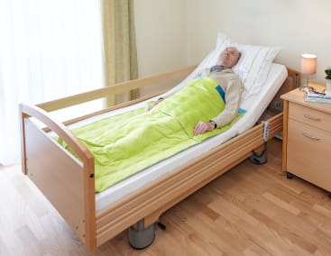 In einem aufgeräumten Zimmer mit hellen Wänden, Holzfußboden, Nachttisch und Lampe liegt ein älterer Mann in einem krankenhausähnlichen Bett mit Holzschienen, zugedeckt mit einer grünen Decke.