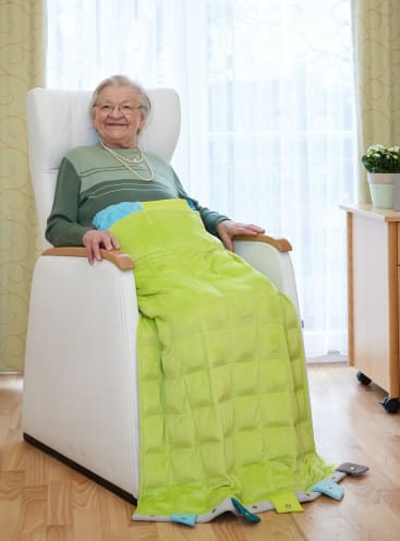 Eine ältere Frau sitzt in einem weißen Sessel mit einer grünen Decke über ihren Beinen und lächelt. Sie trägt eine Brille, einen grünen Pullover und Perlen. Der Raum hat einen hellen Holzfußboden, einen Schreibtisch und eine Topfpflanze neben einem Fenster mit durchsichtigen Vorhängen.