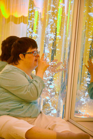 Eine Person, die eine Brille und ein hellblaues Hemd trägt, sitzt auf dem Boden und berührt ein durchsichtiges, beleuchtetes, mit Blasen gefülltes Sinnesrohr, das sie genau betrachtet. Ihr Spiegelbild ist in dem Glas neben ihr zu sehen.