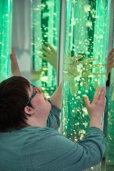 Eine Person mit kurzen Haaren und Brille berührt eine große, vertikale, transparente Röhre, die mit grünem Wasser und schwimmenden Blasen gefüllt ist, deren Spiegelung auf der Oberfläche der Röhre sichtbar ist.