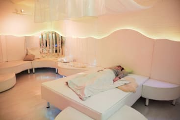 Eine Person liegt auf einem Bett unter einer leichten Decke in einem sanft beleuchteten, weißen Sinnesraum mit geschwungenen Sitzmöbeln, Spiegeln und Hängelampen im Hintergrund.