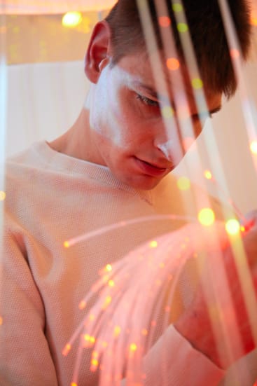 Eine Person, die einen hellen Pullover trägt, blickt genau auf leuchtende Glasfaserstränge mit warmen orangefarbenen und gelben Lichtern. Das Bild ist aus einem Winkel leicht oberhalb der Augenhöhe aufgenommen.