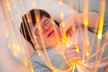 Eine Person mit Down-Syndrom, die eine Brille und ein graues gepunktetes Hemd trägt, liegt auf einer weißen Fläche und hält leuchtende orangefarbene Glasfaserstränge in der Hand, die sie umgeben.