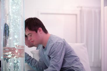 In einem hellen Raum mit weißen Wänden und Vorhängen sitzt ein junger Mensch mit Down-Syndrom auf einem Bett und berührt sanft ein großes, durchsichtiges Rohr, das mit blubberndem Wasser und Lichtern gefüllt ist.